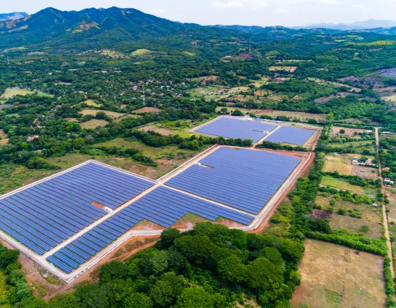 Aerial view of a large solar farm with rows of solar panels on a grassy landscape, surrounded by trees and distant mountains under a partly cloudy sky.