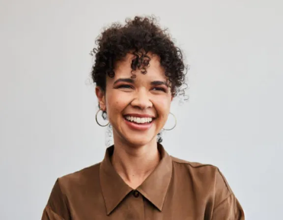 A person with curly hair and hoop earrings smiles while wearing a brown collared shirt against a plain background.