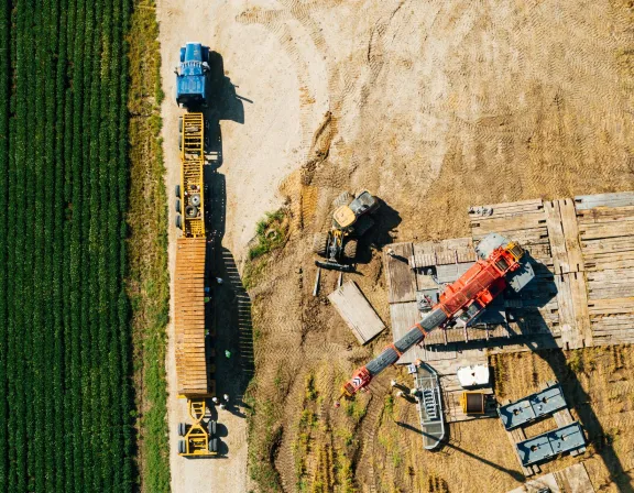 Aerial view of a construction site with a blue truck and a yellow trailer on a dirt road next to a green field. A red crane is positioned on wooden planks with other construction equipment nearby. The ground shows tire tracks and disturbed soil.