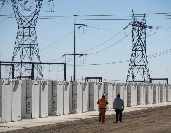 Two workers walk along a row of large, white battery storage units. High-voltage power lines and towers are visible in the background under a clear blue sky.
