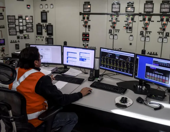 A person in an orange safety vest is sitting at a control desk with multiple computer monitors displaying technical data. The background shows a wall filled with control panels and switches.