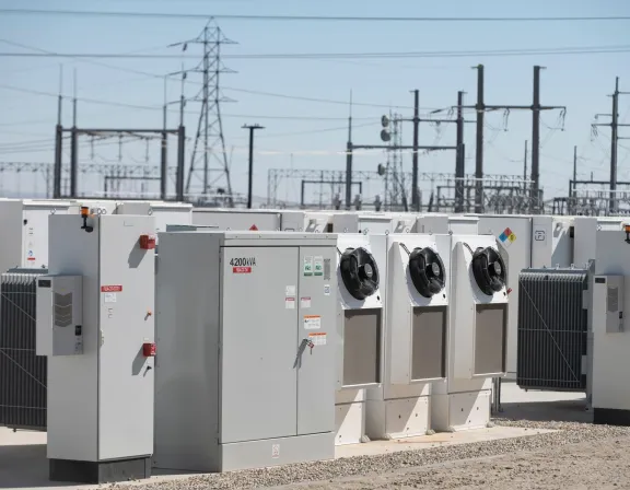 Outdoor electrical transformers and power equipment in a substation with transmission towers in the background.