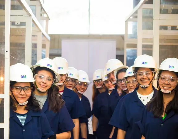 A group of AES women wearing white hard hats and safety goggles stand in two rows inside an industrial setting. They are smiling and wearing dark blue uniforms with the AES logo on their helmets.