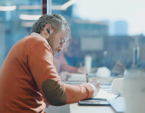 Man in an orange sweater writing in a notebook at a desk in a modern office with headphones in his ears.