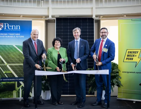 Four individuals in formal attire participate in a ribbon-cutting ceremony. They stand in front of banners for the University of Pennsylvania and Energy Week, with solar panels in the background.
