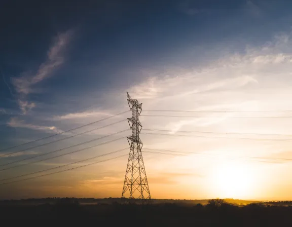 A tall electricity pylon stands against a colorful sunset sky with scattered clouds and power lines stretching across the scene.