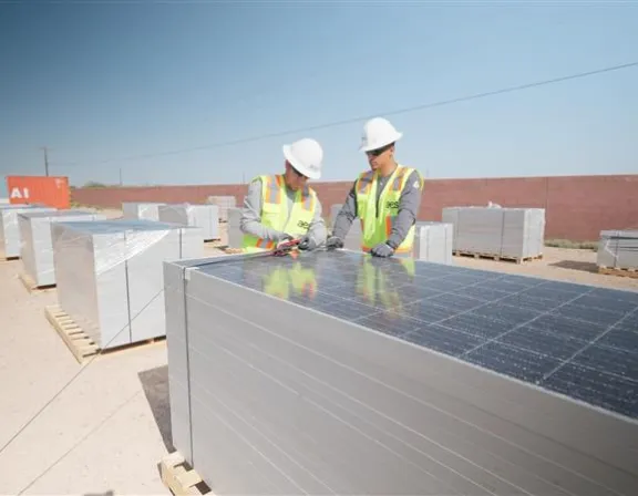 Two workers in safety gear inspecting solar panels stacked on pallets in an outdoor area with clear skies.