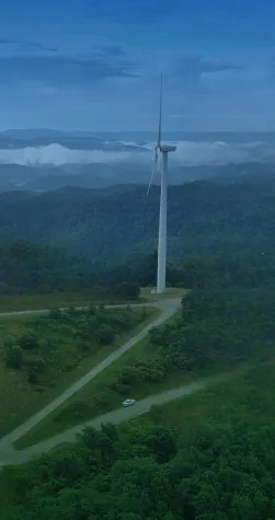 Aerial view of a wind farm with multiple wind turbines on a lush green hilltop, surrounded by dense forests and distant mountain ranges under a clear blue sky.