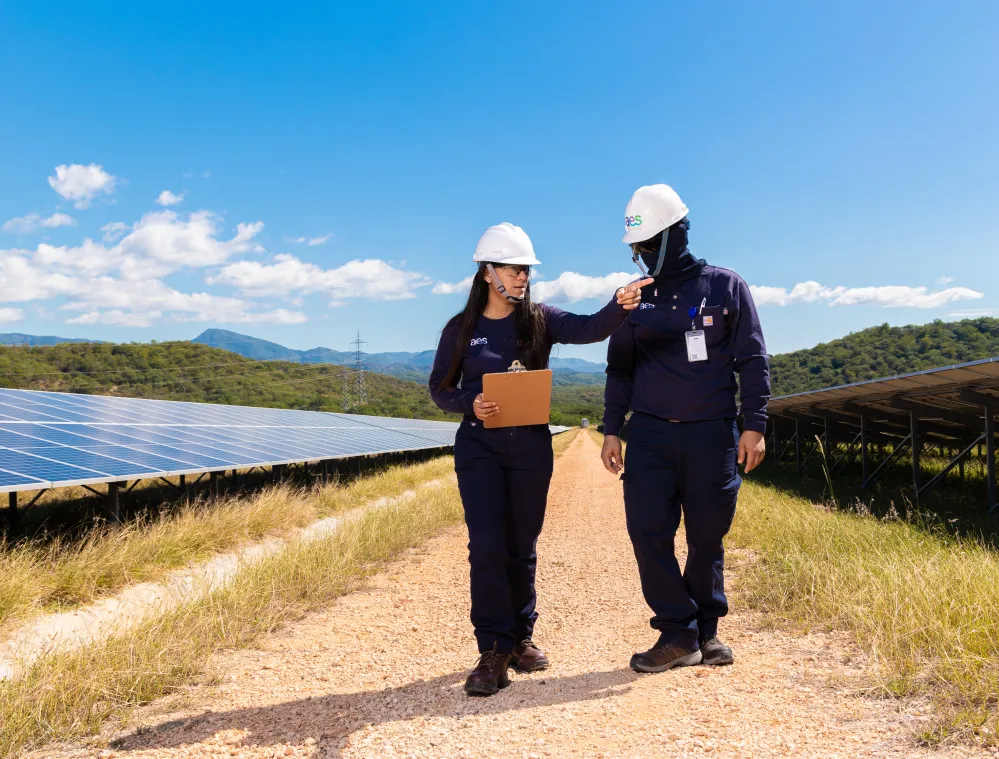 Two workers in protective gear and helmets walking beside solar panels in a sunny field, discussing and holding a clipboard.