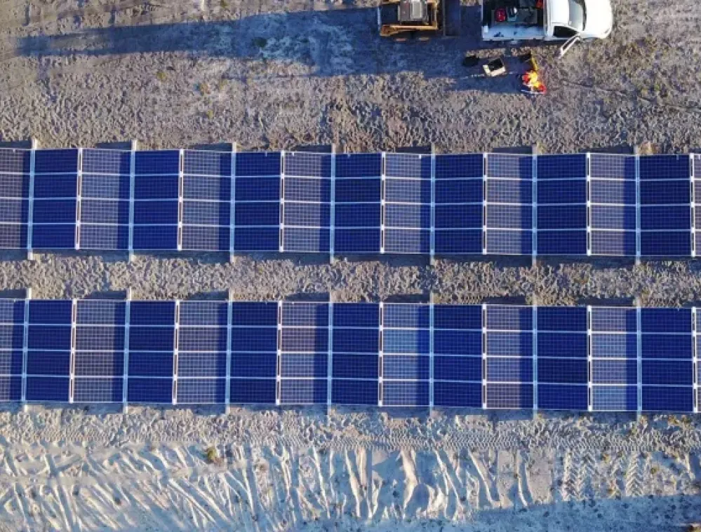 Aerial view of two rows of solar panels installed on a sandy surface. A white vehicle and some equipment are parked nearby, with two people wearing orange safety gear standing beside the vehicle.