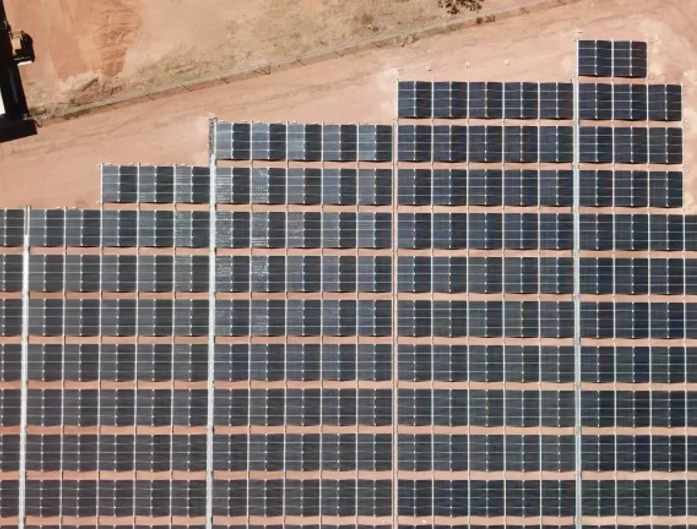 Aerial view of a large solar panel array arranged in a grid pattern on a sandy terrain. The panels are aligned in rows and columns, covering a significant portion of the ground.