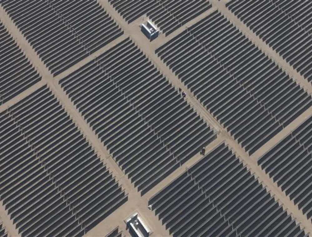 Aerial view of a large solar panel farm on a sandy terrain, with rows of dark solar panels arranged in a grid pattern.