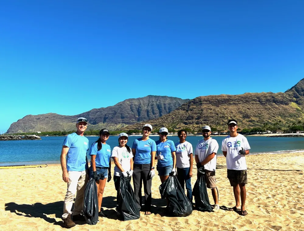 A group of eight people stands on a sandy beach holding black trash bags. They are participating in a cleanup activity under a clear blue sky, with mountains and a body of water in the background.