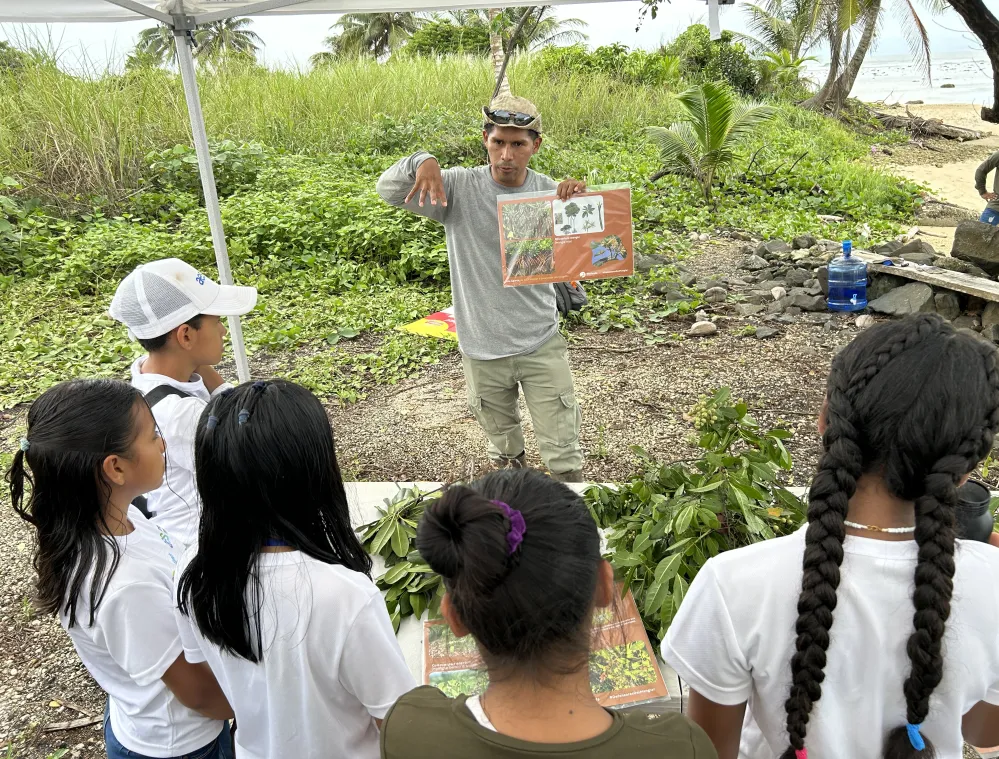A group of children in white shirts listen to a man holding educational posters, standing under a canopy near a beach. The man is gesturing while explaining, surrounded by greenery and palm trees.