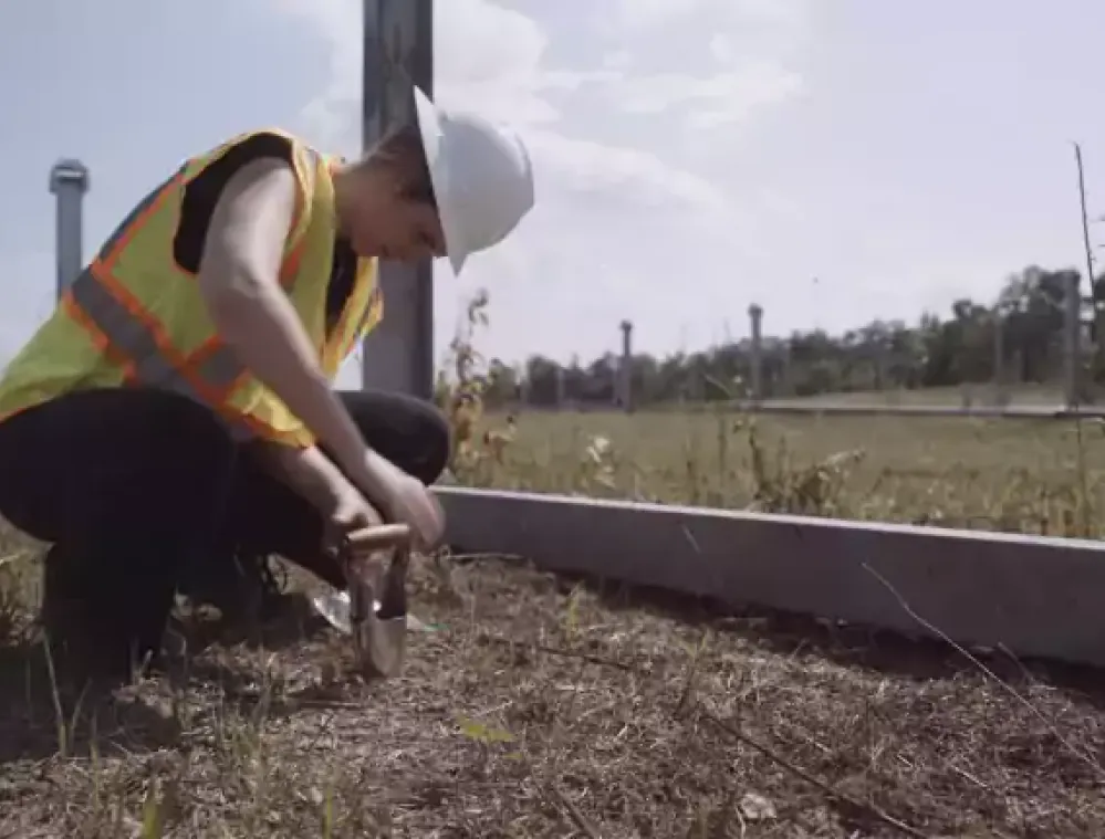 A person wearing a white hard hat and a reflective vest crouches down to take soil samples in a grassy area near a road. The background shows a clear sky and trees.