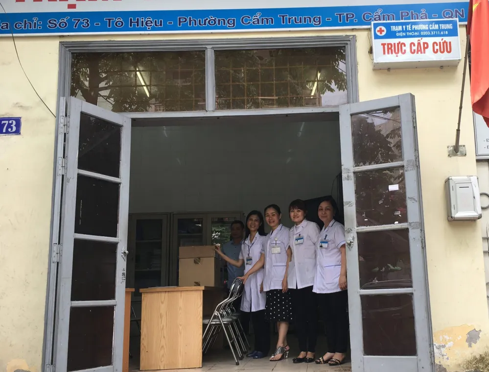 A group of five medical staff in white coats stand at the entrance of a healthcare center. The building has a sign in Vietnamese above the door. The entrance is open, revealing a room with chairs and a desk.