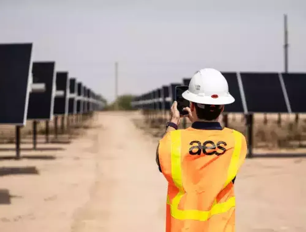 A person wearing a hard hat and reflective vest stands in a solar farm, taking a photo of rows of solar panels.
