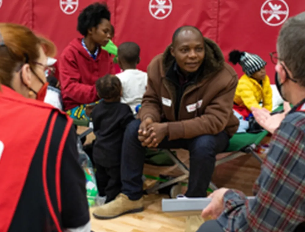 A group of people sitting on cots in a shelter, with two individuals wearing Red Cross vests talking to them. Children are present, and a red backdrop with Red Cross symbols is visible.