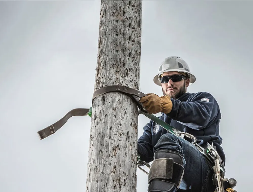A utility worker wearing a hard hat and safety gear climbs a wooden pole using a harness and climbing equipment.