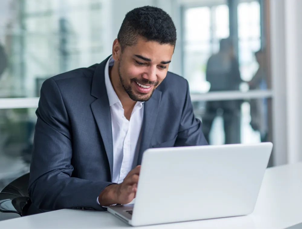 A man in a suit smiling while working on a laptop at a desk in a modern office.