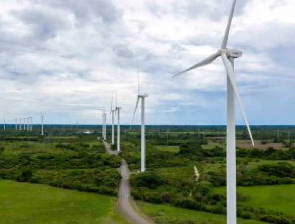 A row of wind turbines in a green field under a cloudy sky. The turbines are aligned along a dirt path, stretching into the distance.