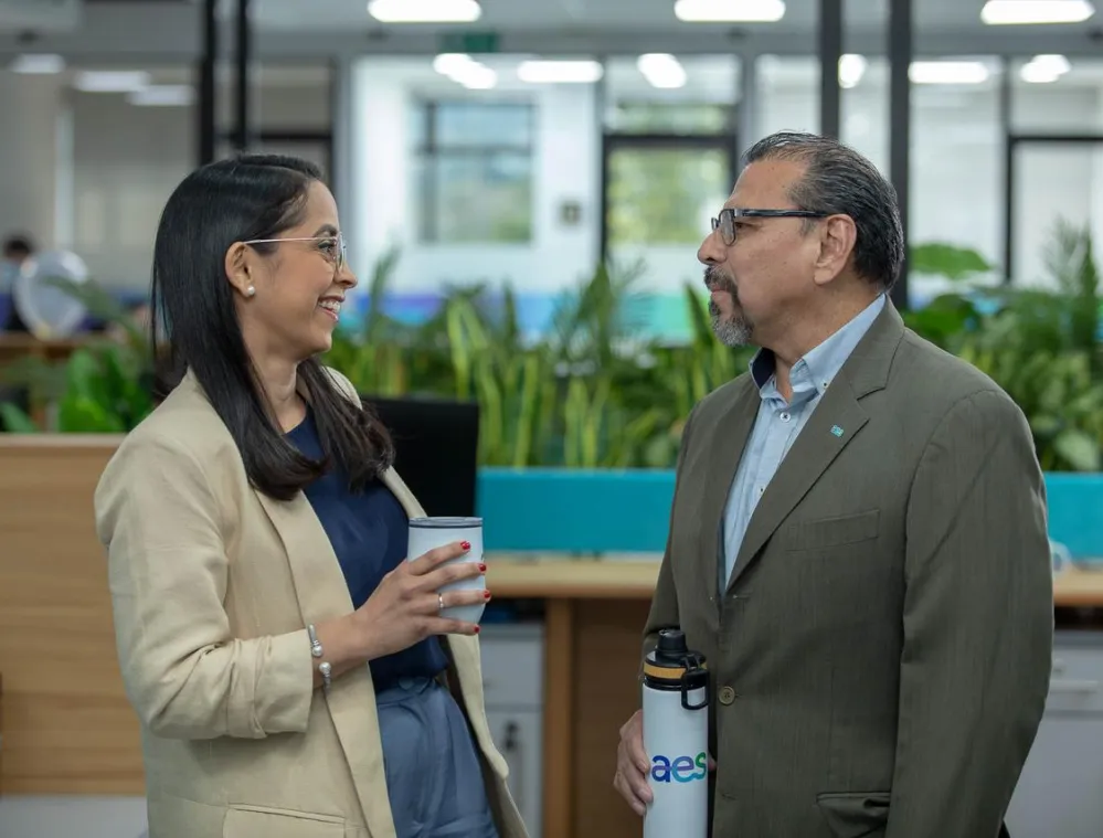 Two colleagues in an office setting, a woman in a beige blazer holding a coffee cup, and a man in a green suit with a water bottle, engaging in conversation.