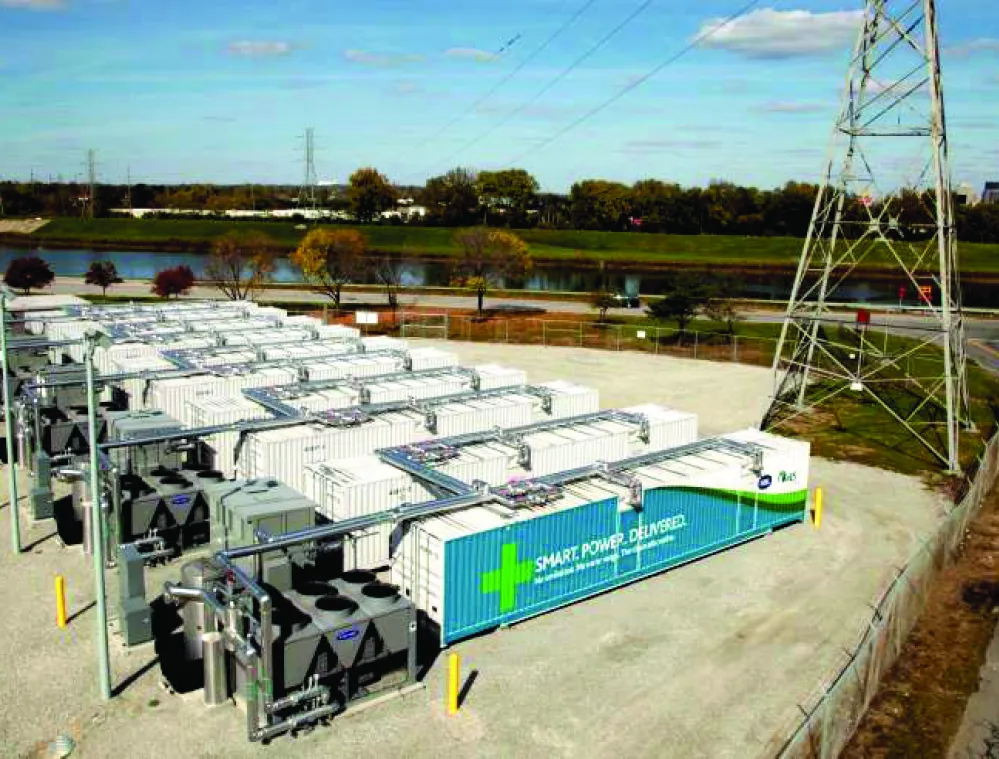 Aerial view of a power storage facility with multiple white containers and electrical equipment. A tall metal transmission tower stands nearby, and a road curves around the facility. Trees and a river are visible in the background under a partly cloudy sky.