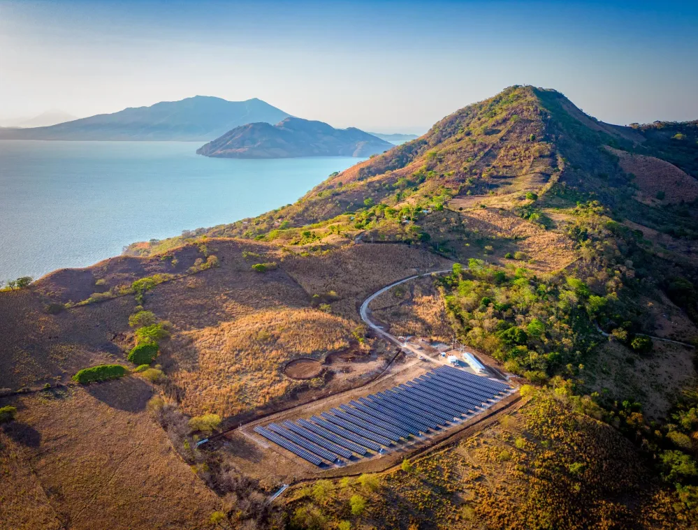 Aerial view of a solar farm nestled in a hilly landscape near a large body of water, with mountains visible in the background under a clear blue sky.