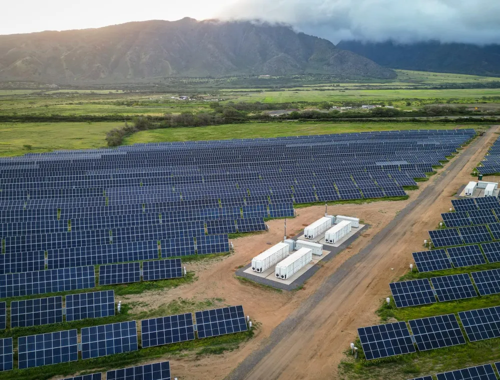 Aerial view of a large solar farm with rows of solar panels and several white utility buildings. The farm is set in a green landscape with mountains in the background under a cloudy sky.