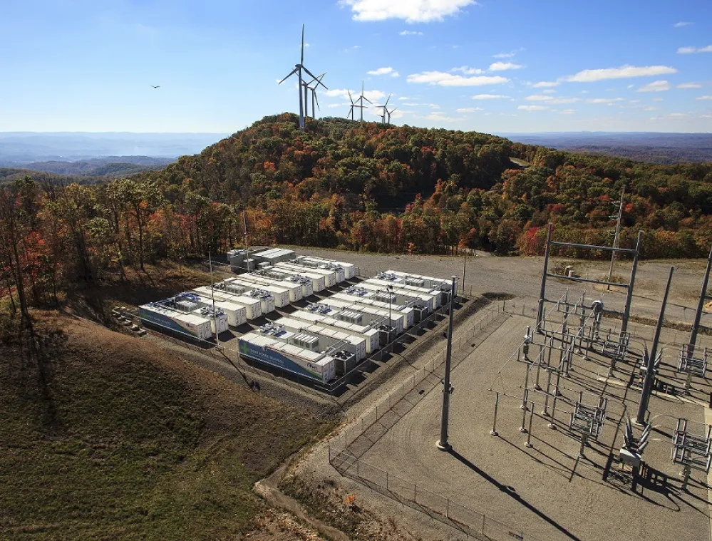 Aerial view of a wind farm and energy storage facility on a hill with several wind turbines and battery containers, surrounded by autumn-colored forest.
