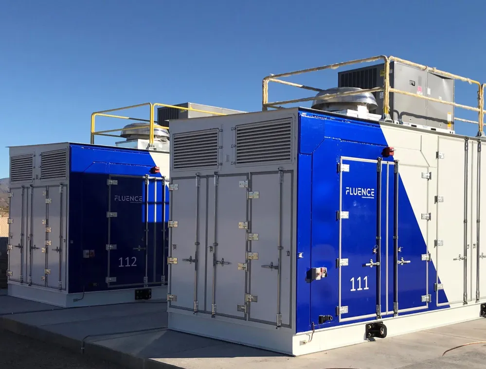 Two large industrial battery storage units with blue and white panels, labeled Fluence, are situated outdoors under a clear blue sky.