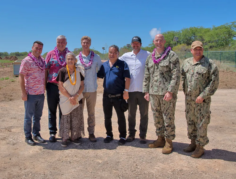A group of eight people standing outdoors on a sunny day, some wearing leis and military uniforms, smiling at the camera with a clear sky and trees in the background.