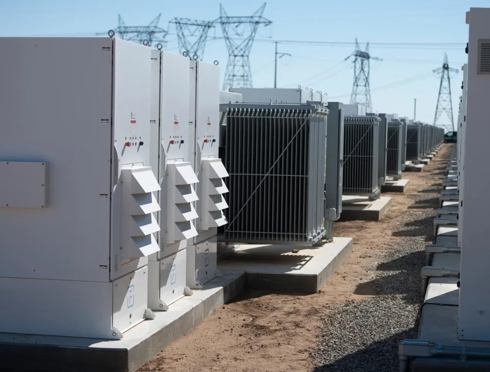 A row of large electrical transformers and equipment is aligned on concrete platforms outdoors. Power lines and towers are visible in the background under a clear blue sky.