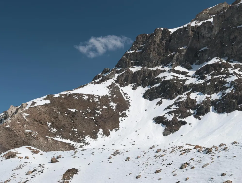 Snow-covered mountain slope with rocky outcrops under a clear blue sky. Sparse patches of snow reveal brown earth and rocks.