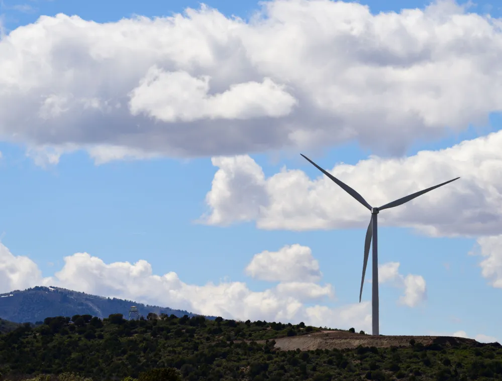 A wind turbine stands on a hill under a blue sky with scattered white clouds. A mountain range is visible in the background.