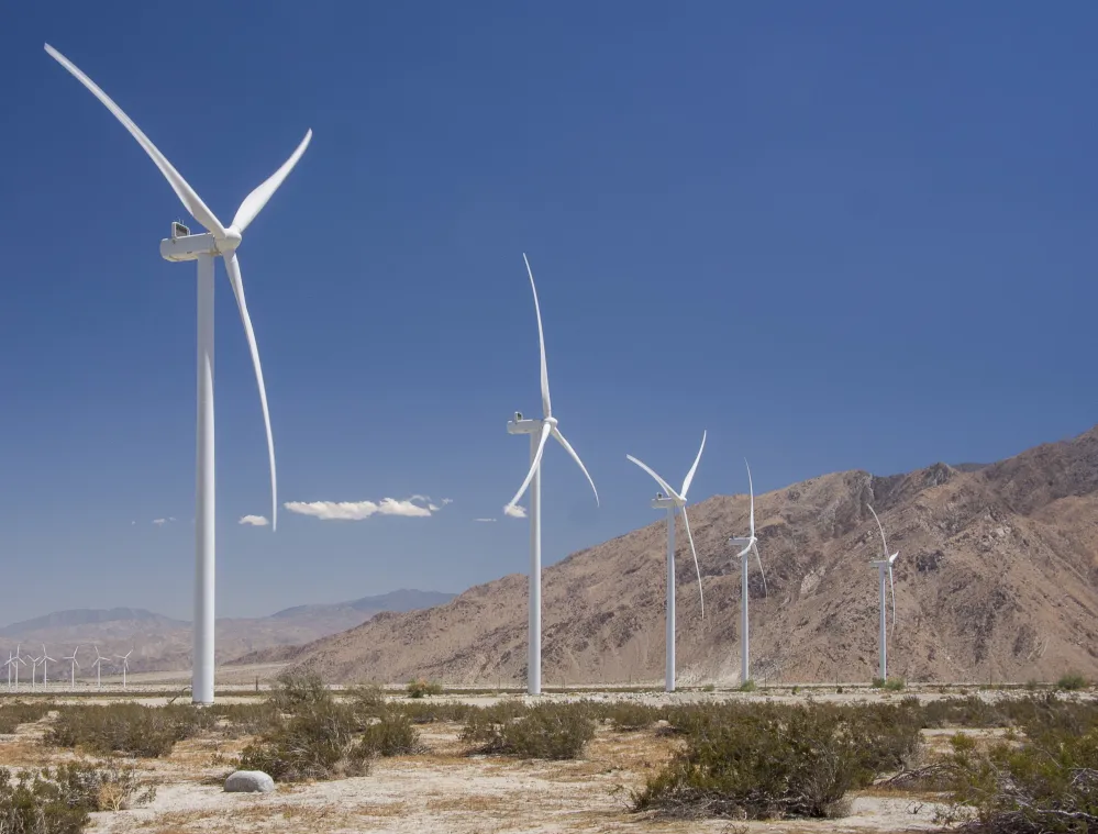 A row of white wind turbines in a desert landscape with mountains in the background under a clear blue sky.