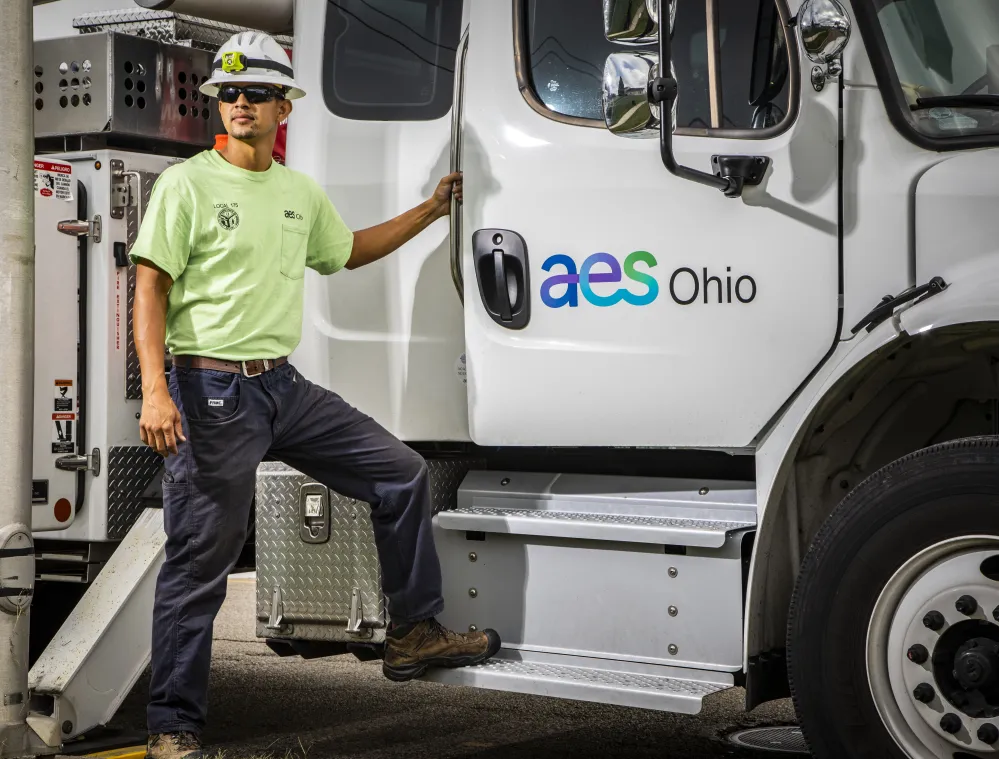 A worker in a green shirt and hard hat stands next to a white truck with 'aes Ohio' logo, holding the door handle.