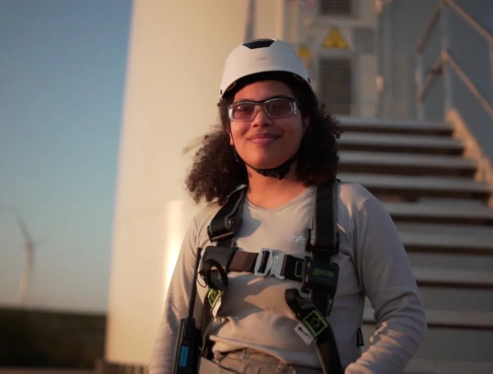 A person wearing safety gear, including a helmet and harness, stands in front of a wind turbine with stairs. Wind turbines are visible in the background under a clear sky.