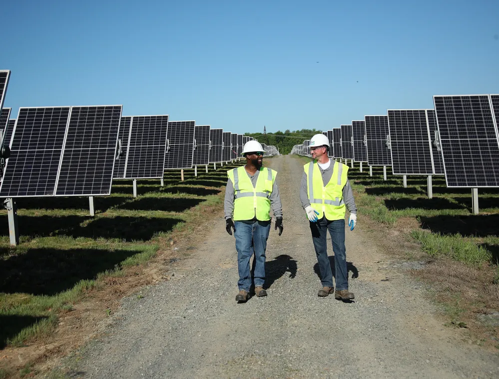 Two workers in safety vests and helmets walk along a path between rows of solar panels under a clear blue sky.