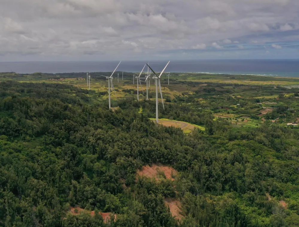 Aerial view of a lush green landscape with several wind turbines scattered across the area. The turbines are surrounded by dense forests and open fields. In the background, the ocean stretches under a cloudy sky.