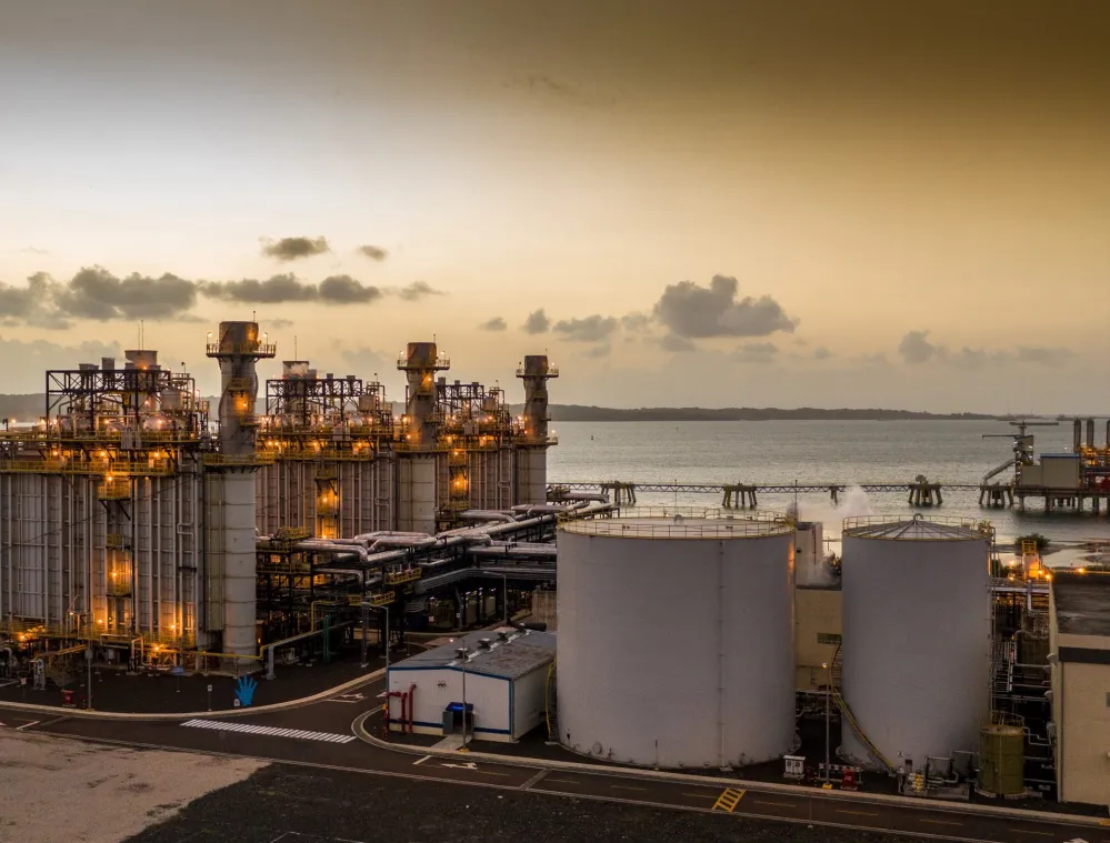 An industrial facility with large storage tanks and complex piping systems is illuminated at dusk. The facility is situated near a body of water, with a long pier extending into the distance. The sky is overcast with scattered clouds.