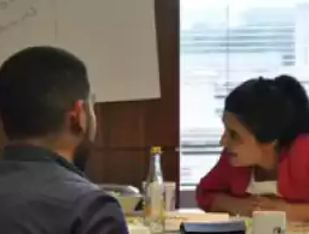 Four people sit around a table engaged in a discussion in an office setting, with papers, cups, and a bottle on the table and a whiteboard in the background.