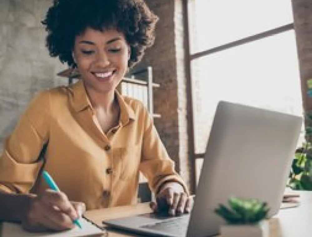 Smiling woman sitting at a desk, writing in a notebook while using a laptop in a bright, modern office with a large window and a small potted plant.