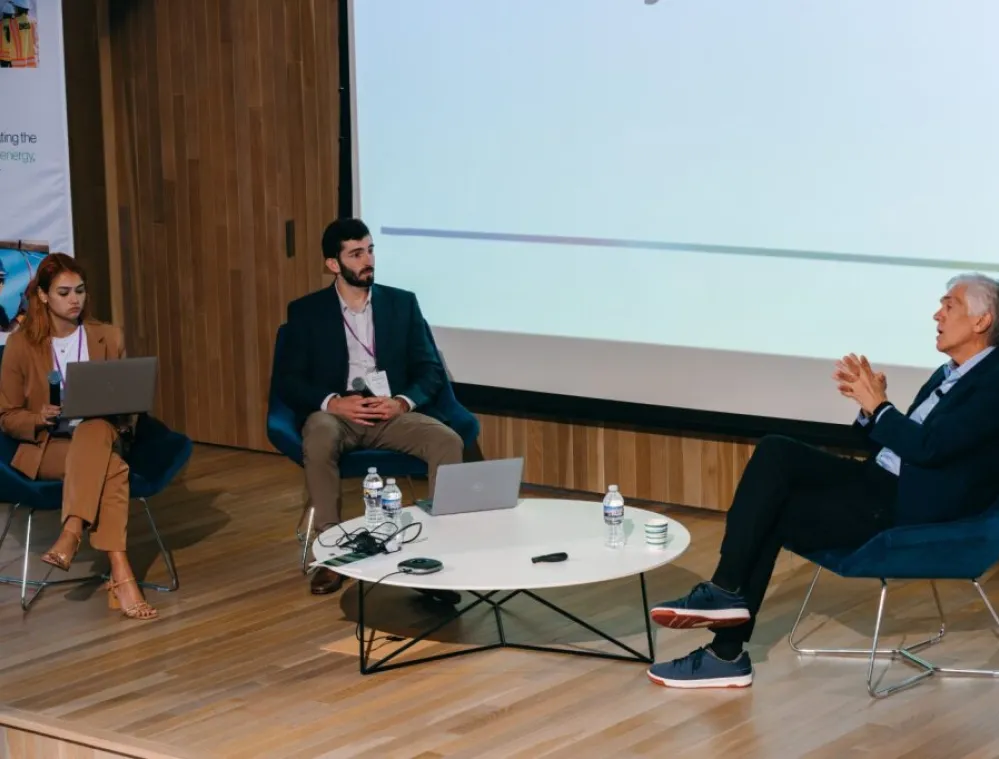 Three people sit in modern chairs on a stage during a panel discussion. Two participants have laptops open and one is speaking. A round table with water bottles, a microphone, and cables is in the center. A large screen and a banner about energy are visible in the background.