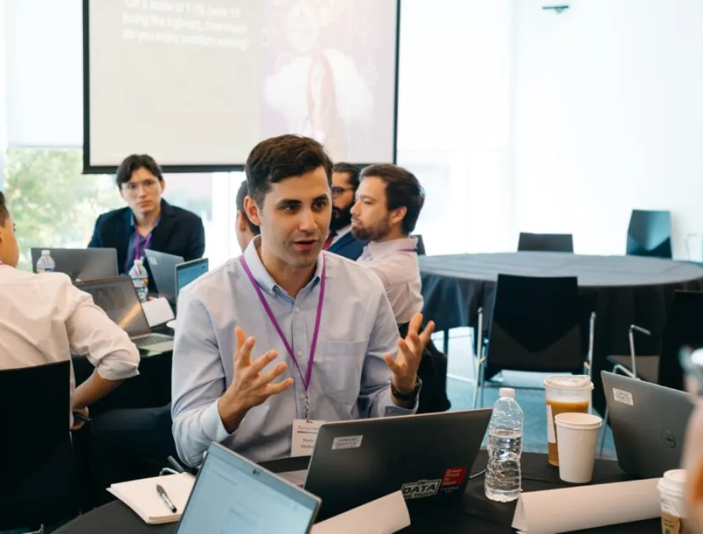 A group of professionals engaged in a discussion around a conference table with laptops, coffee cups, and water bottles. One man in the foreground is speaking and gesturing with his hands. A presentation screen is visible in the background.