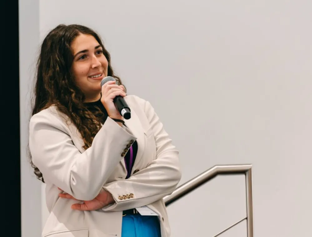 A woman with long curly hair wearing a white blazer stands indoors, holding a microphone and smiling while speaking. She is positioned near a metal railing and a plain light-colored wall.