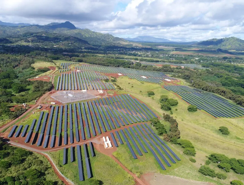 Aerial view of a large solar farm with rows of solar panels spread across a green landscape. The panels are surrounded by trees and grass, with mountains visible in the background under a partly cloudy sky.