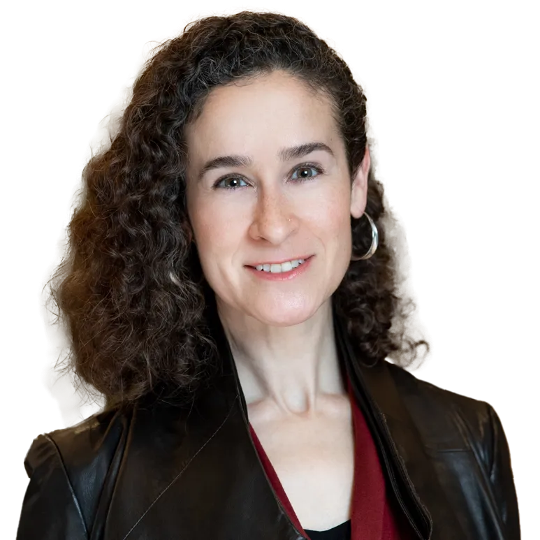 Smiling person with curly hair and hoop earrings wearing a leather jacket, standing indoors with a blurred background.