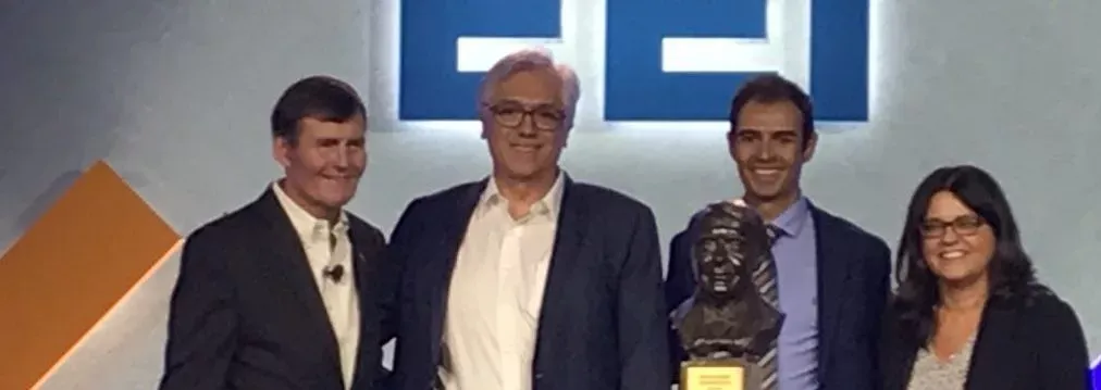 Four people standing together at an event, three men and one woman, with one man holding a sculpture. A logo is visible in the background.