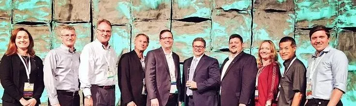 A group of ten people standing in a row, dressed in business attire, smiling in front of a textured wall with a greenish hue.